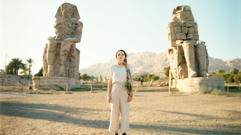 A woman standing in front of the Colossi of Memnon, two massive ancient stone statues, with a bright blue sky in the background