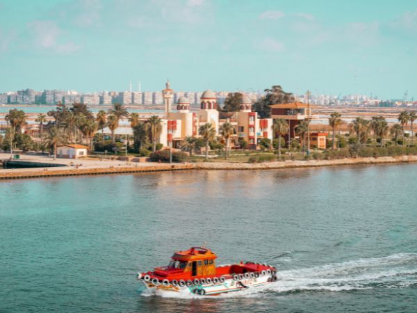 A vibrant red boat cruising along a calm river, with palm trees, buildings, and a cityscape in the background