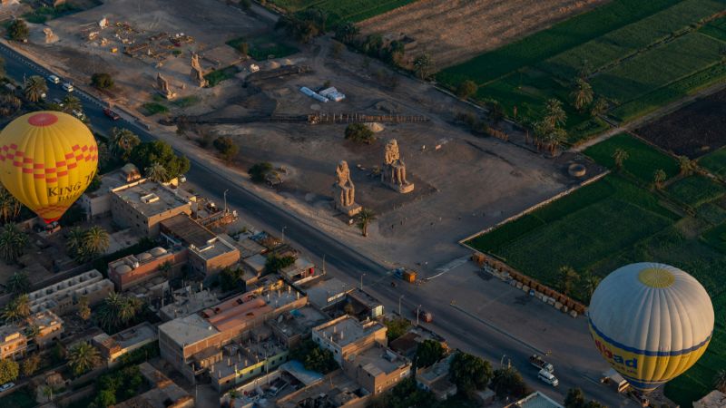 An aerial view of the Colossi of Memnon and surrounding fields, with colorful hot air balloons floating above the landscape