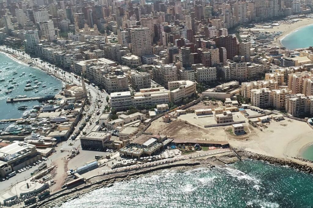 A scenic view of Alexandria's waterfront with Mediterranean waves, historic architecture, and palm trees under a bright sky.