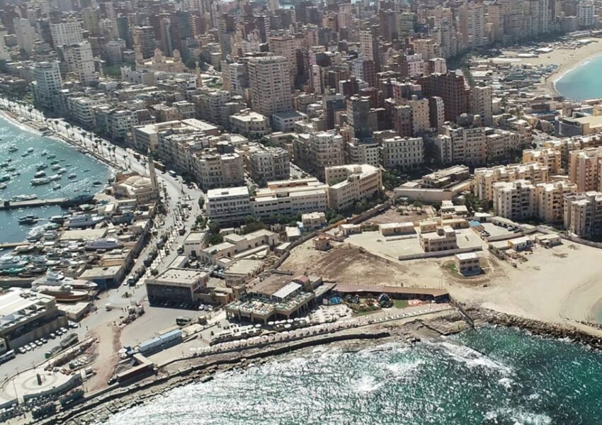 A scenic view of Alexandria's waterfront with Mediterranean waves, historic architecture, and palm trees under a bright sky.