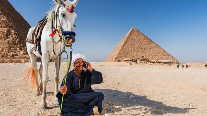 Local man sitting near the pyramids with a mobile phone and a horse