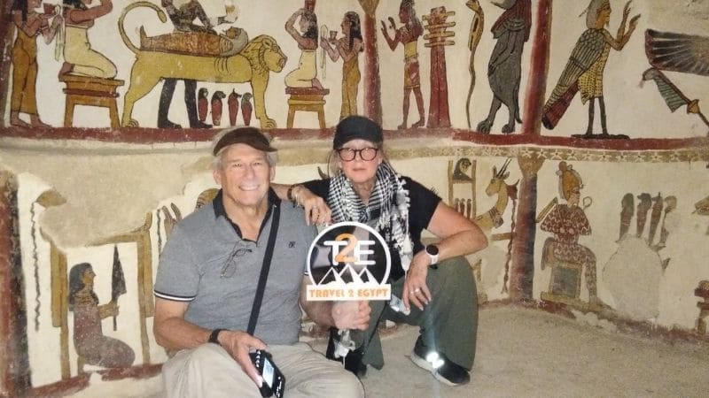 Two tourists inside an ancient tomb, surrounded by colorful wall paintings and hieroglyphics