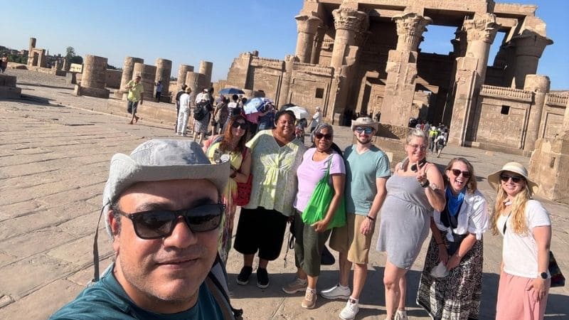 A group of tourists posing in front of the Temple of Kom Ombo, with stone columns and the temple façade visible