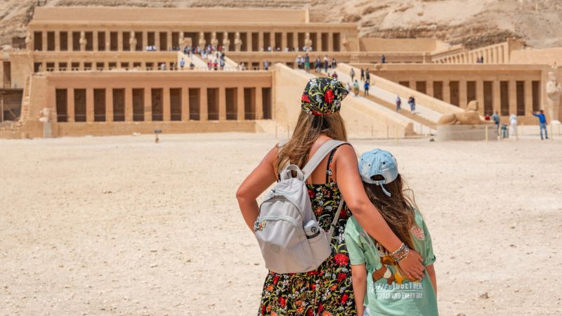 A mother and child standing in front of the grand Temple of Hatshepsut at Deir el-Bahari, surrounded by a desert landscape