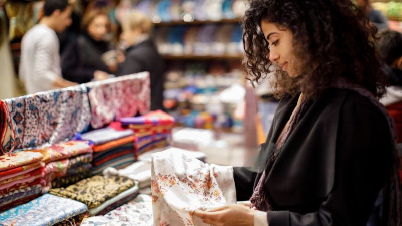 A woman examining colorful fabrics at a market, with other shoppers in the background