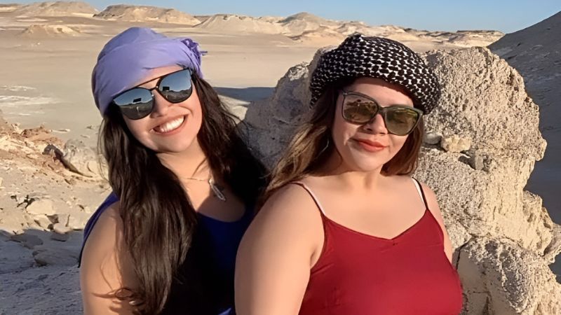 Two women wearing headscarves and sunglasses smiling in a desert landscape