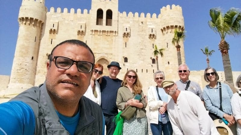 A group of tourists posing in front of the Citadel of Qaitbay in Alexandria, Egypt, under a clear blue sky