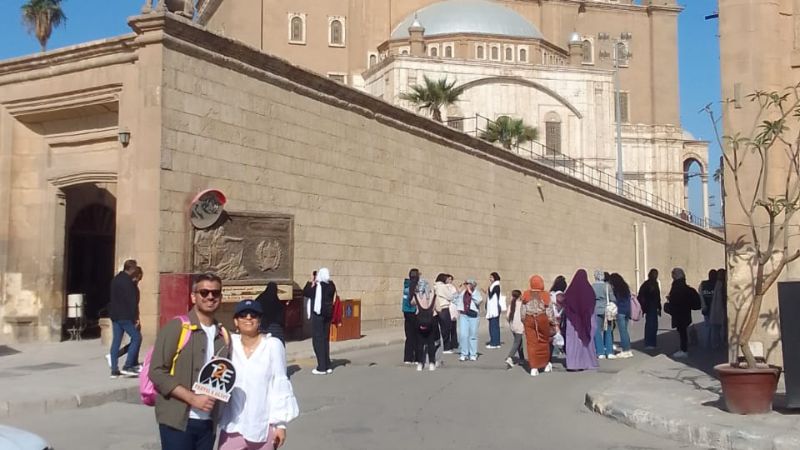 A couple posing near a historic Egyptian church with a group of tourists in the background