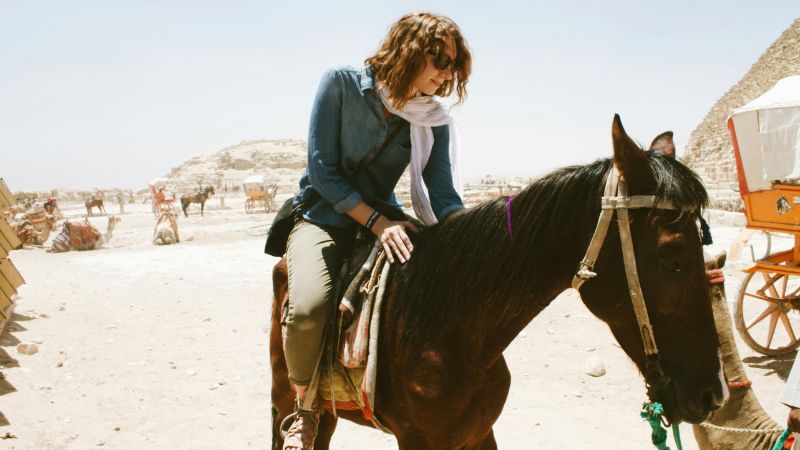 A woman riding a horse near the Pyramids of Giza on a sunny day, dressed casually with a scarf around her neck