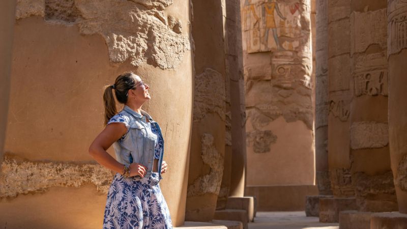 A tourist admiring the massive sandstone columns of Karnak Temple, with intricately carved hieroglyphs glowing in the sunlight