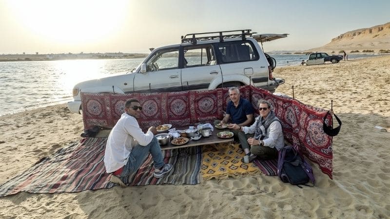 A tranquil lakeside dining scene in Egypt’s desert, with a beautifully set table overlooking a serene desert lake under a pastel-colored sky