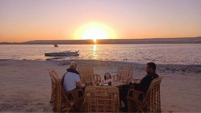 A serene lakeside scene in Egypt at sunset, with calm waters reflecting the golden and pink hues of the sky, surrounded by desert terrain