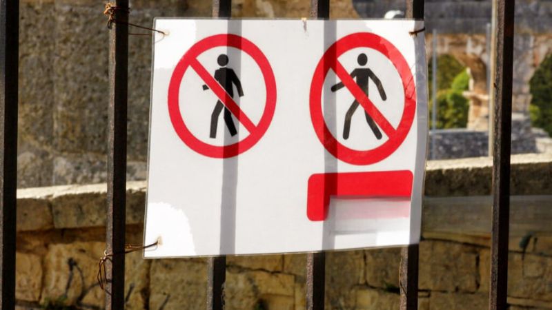 A sign with two red prohibition symbols, indicating no walking or climbing, attached to a metal fence with a historic structure in the background