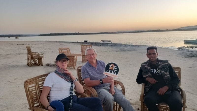 A peaceful lakeshore scene in Egypt at sunset, with a traveler seated by the calm waters, enjoying the golden and pink hues of the sky