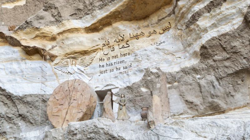 A carved stone wall with biblical inscriptions and a depiction of Jesus' resurrection in a cave-like setting