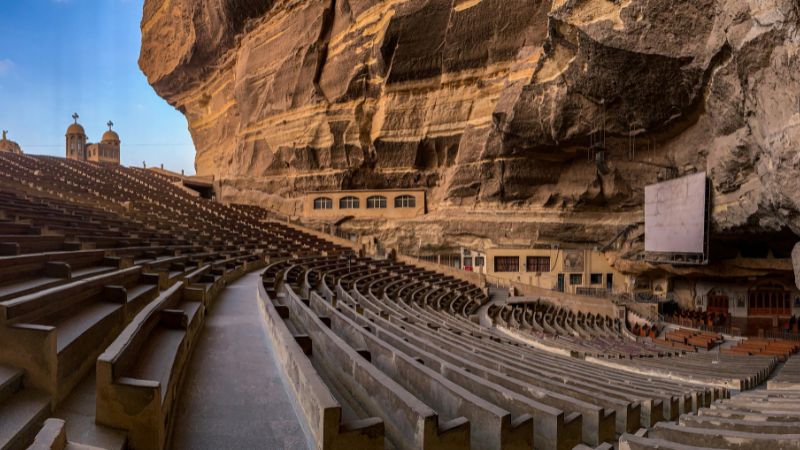 A vast open-air amphitheater carved into a rock formation with seating rows and a stunning natural backdrop