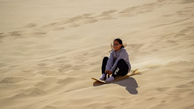 A young girl sandboarding down a dune in a desert