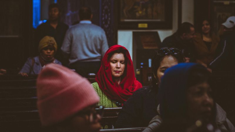 A woman in a red scarf meditating or praying inside a church, surrounded by other worshippers