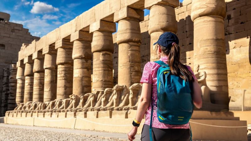 A traveler walking toward the Avenue of Ram-Headed Sphinxes at Karnak Temple, under a bright and sunny sky