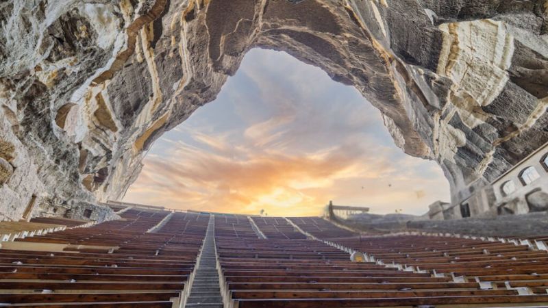 A view of a rock-carved theater with rows of seats facing a dramatic cave opening at sunset