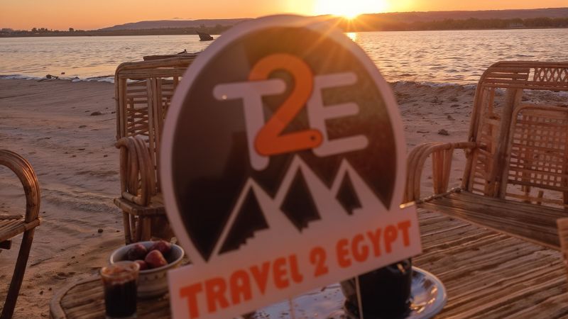 Travel & Egypt" sign placed on a table near the water, with a beautiful sunset in the background