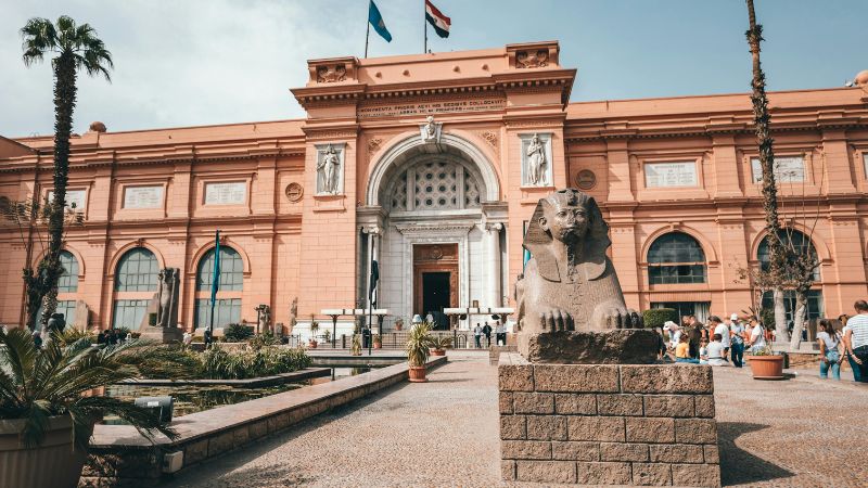 The exterior of the Egyptian Museum in Cairo, featuring a pink-hued neoclassical facade and a seated sphinx statue in the foreground