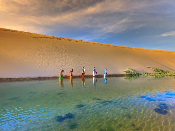 A group of people walking along a calm body of water beside a golden sand dune under a vibrant sky