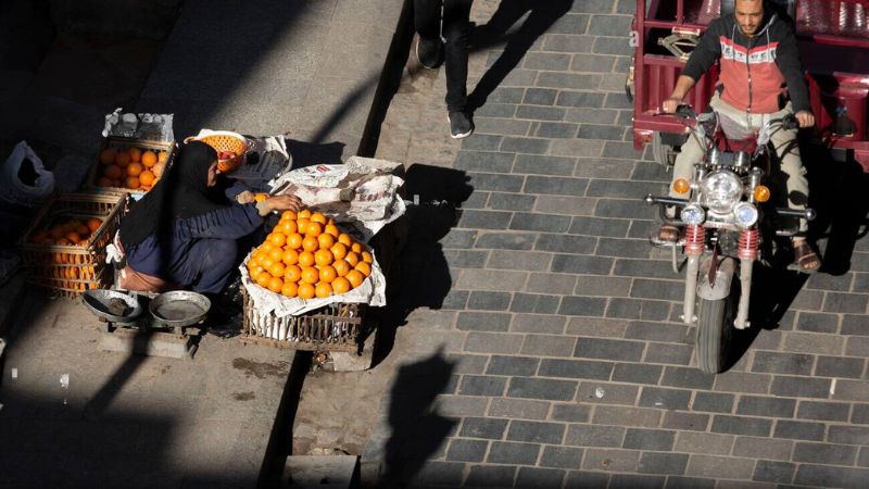 A street vendor arranging oranges on a cart, with a motorbike passing by on a cobbled street