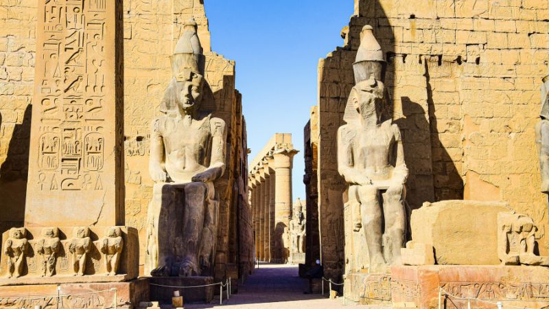 The grand entrance of Luxor Temple, flanked by towering statues of seated pharaohs and lined with ancient columns under a clear blue sky