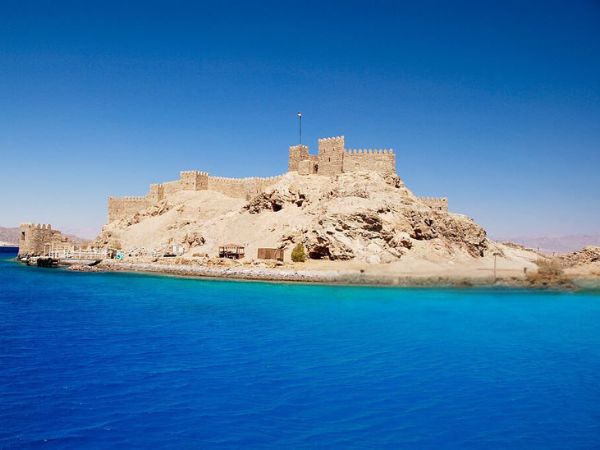 An ancient stone fortress perched on a rocky hill overlooking vivid blue waters under a bright sky