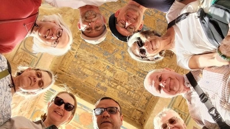 A tour group stands beneath intricately decorated ancient Egyptian ceilings, adorned with colorful hieroglyphs and symbols