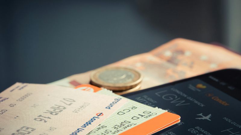 A close-up of travel essentials, including boarding passes, a smartphone displaying flight details, Euro banknotes, and coins