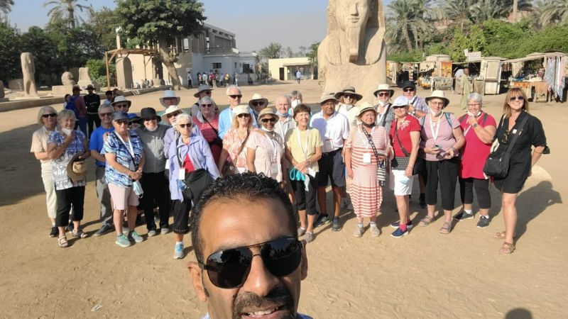 A group of tourists posing near the Great Sphinx of Giza under a sunny sky, surrounded by sand and ancient ruins