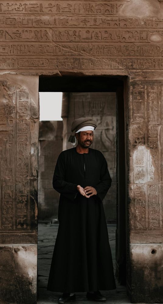 A man wearing a traditional galabeya standing at the entrance of a historical Egyptian site with hieroglyphic carvings