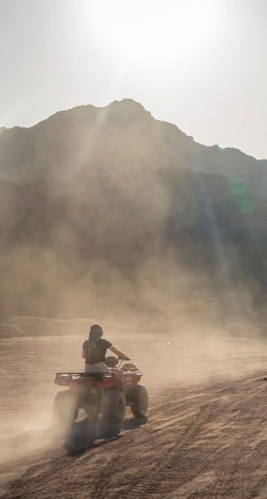 Person riding a quad bike on a dusty desert trail with mountains in the background under the bright sun