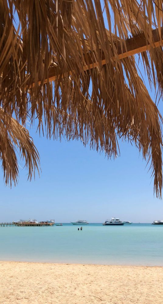 Sandy beach with a thatched roof shade overlooking turquoise waters and anchored boats