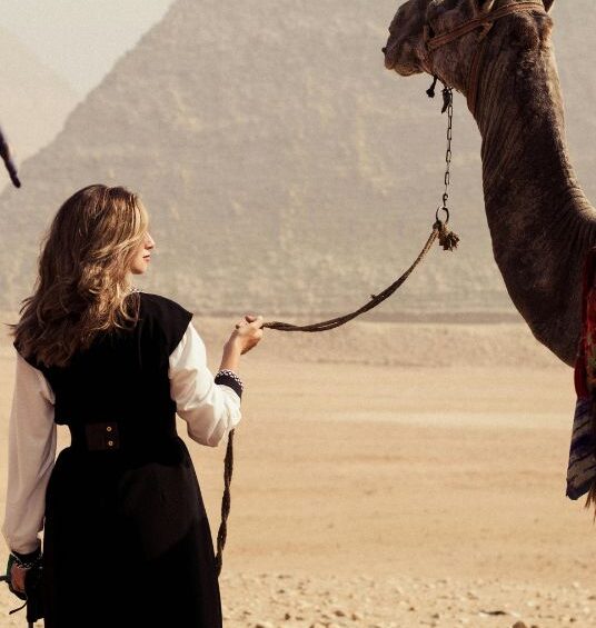 Woman standing with a camel in the desert with the Pyramids of Giza visible in the background