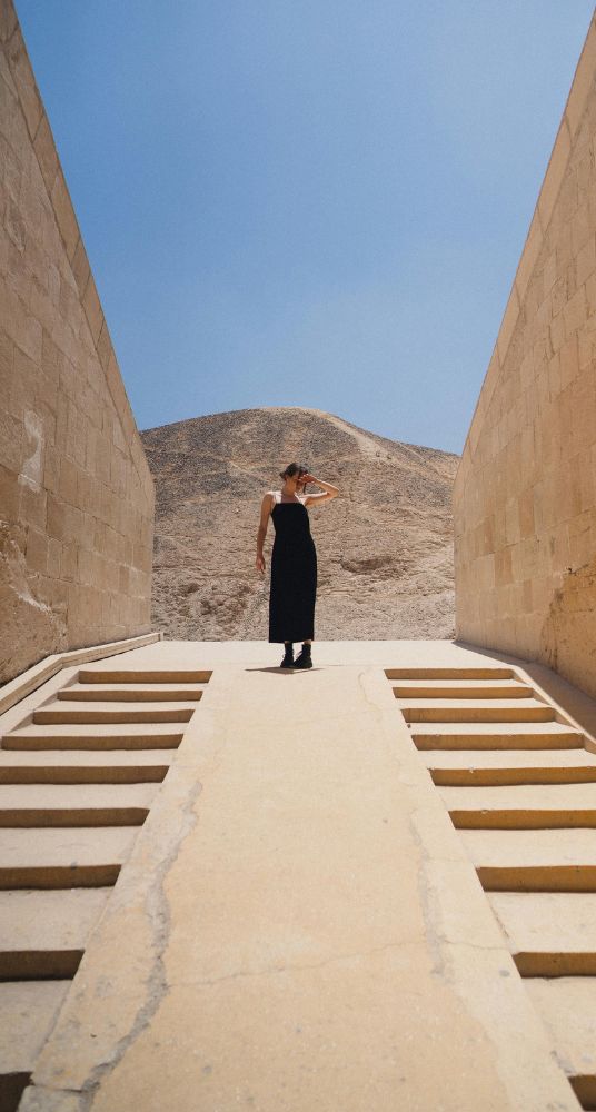 Woman standing between ancient stone walls with a mountainous backdrop under a bright sky