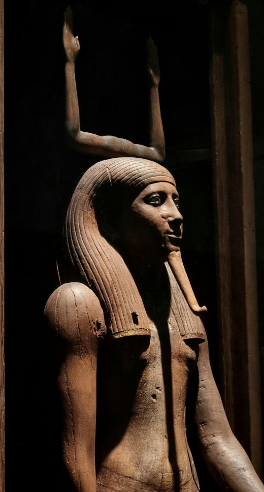 Ancient Egyptian wooden statue with intricate details and raised arms in a museum display