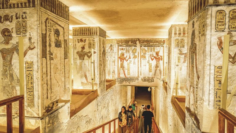 Visitors descend into a tomb in the Valley of the Kings, surrounded by vivid ancient Egyptian wall paintings and carvings.