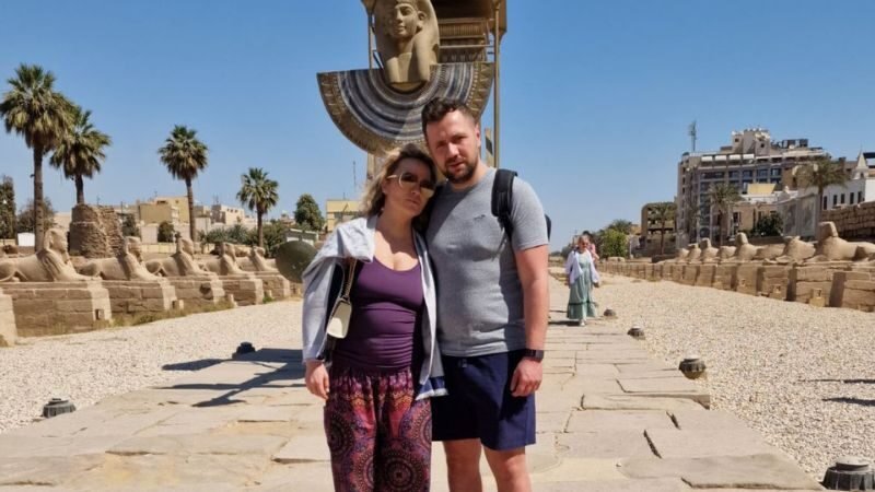 A couple posing at the Avenue of Sphinxes in Luxor Egypt with rows of sphinx statues and a large decorative sphinx monument in the background