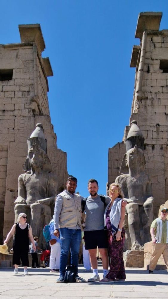 Tourists posing with a guide in front of the entrance to Luxor Temple with colossal statues of Pharaoh Ramses II and ancient Egyptian carvings in the background