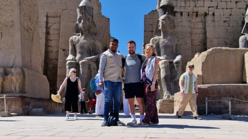 A couple posing with their guide in front of the entrance to Luxor Temple with colossal statues of Pharaoh Ramses II and ancient Egyptian carvings